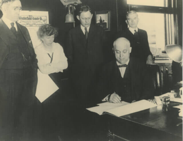 Black and White photo of people in a room while an older man, sitting, signs a document.